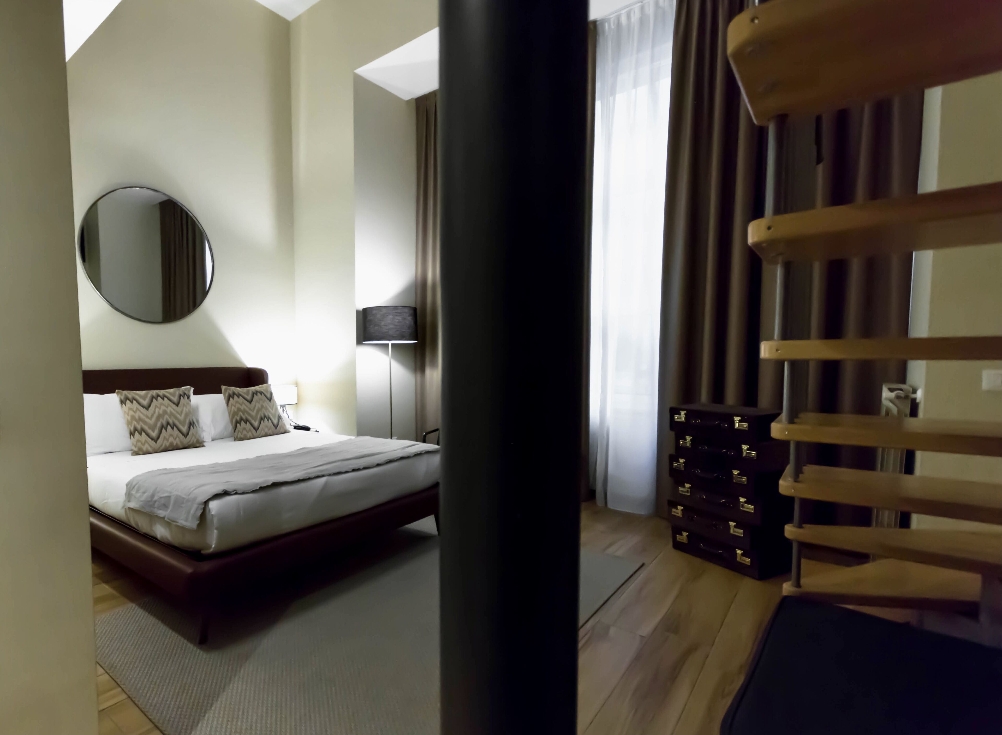 Une chambre d’hôtel avec un lit, un miroir rond, une lampe de chevet, une commode et une vue sur l’extérieur par une fenêtre.