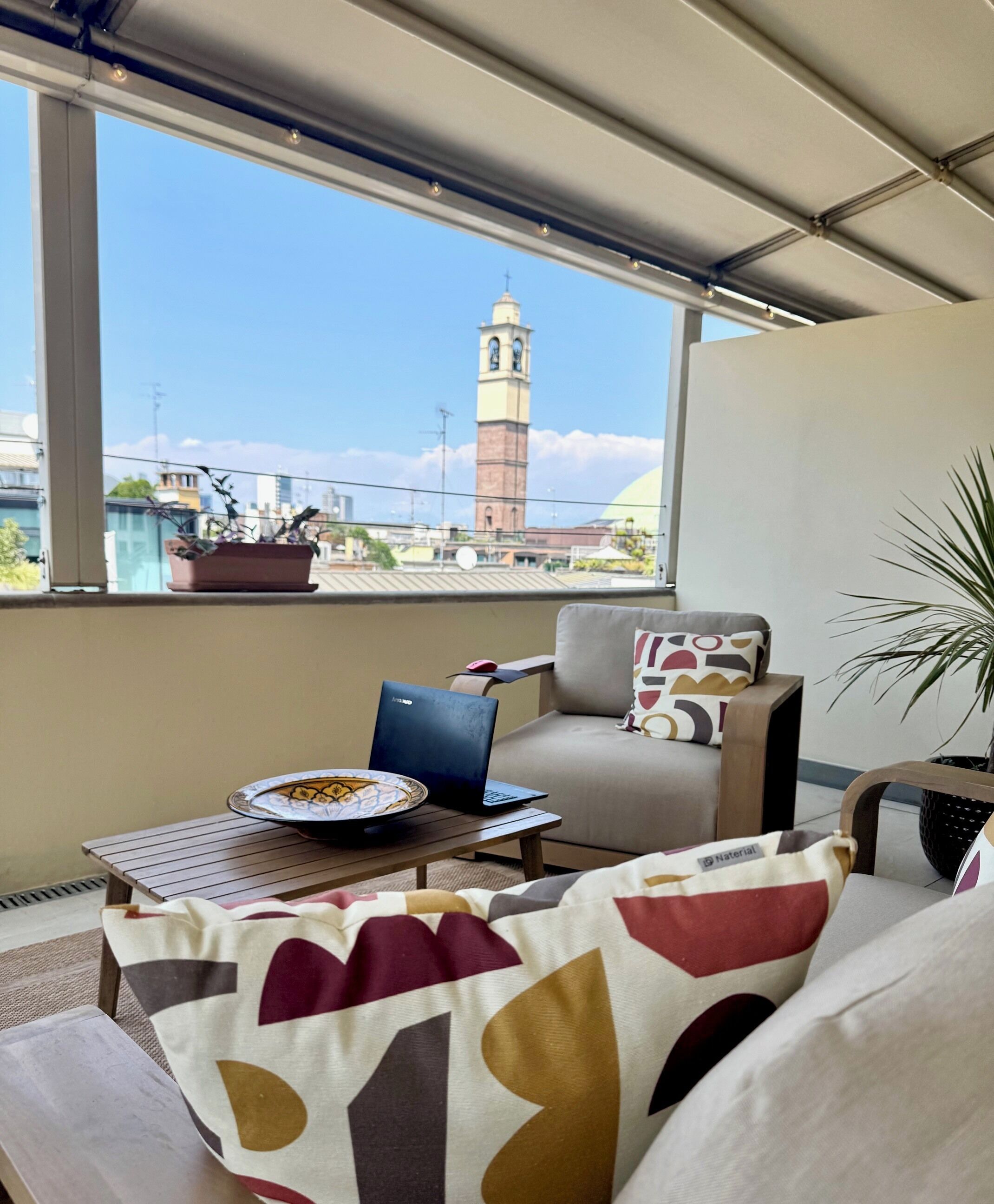 A balcony with a view of a clock tower, a laptop on a table, and a sofa with patterned cushions.