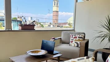 A balcony with a view of a clock tower, a laptop on a table, and a sofa with patterned cushions.