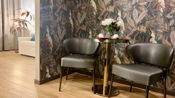 Et værelse med to grå polstrede lænestole omkring et lille rundt bord med guld detaljer, blomstret tapet og en hvid sofa i baggrunden.