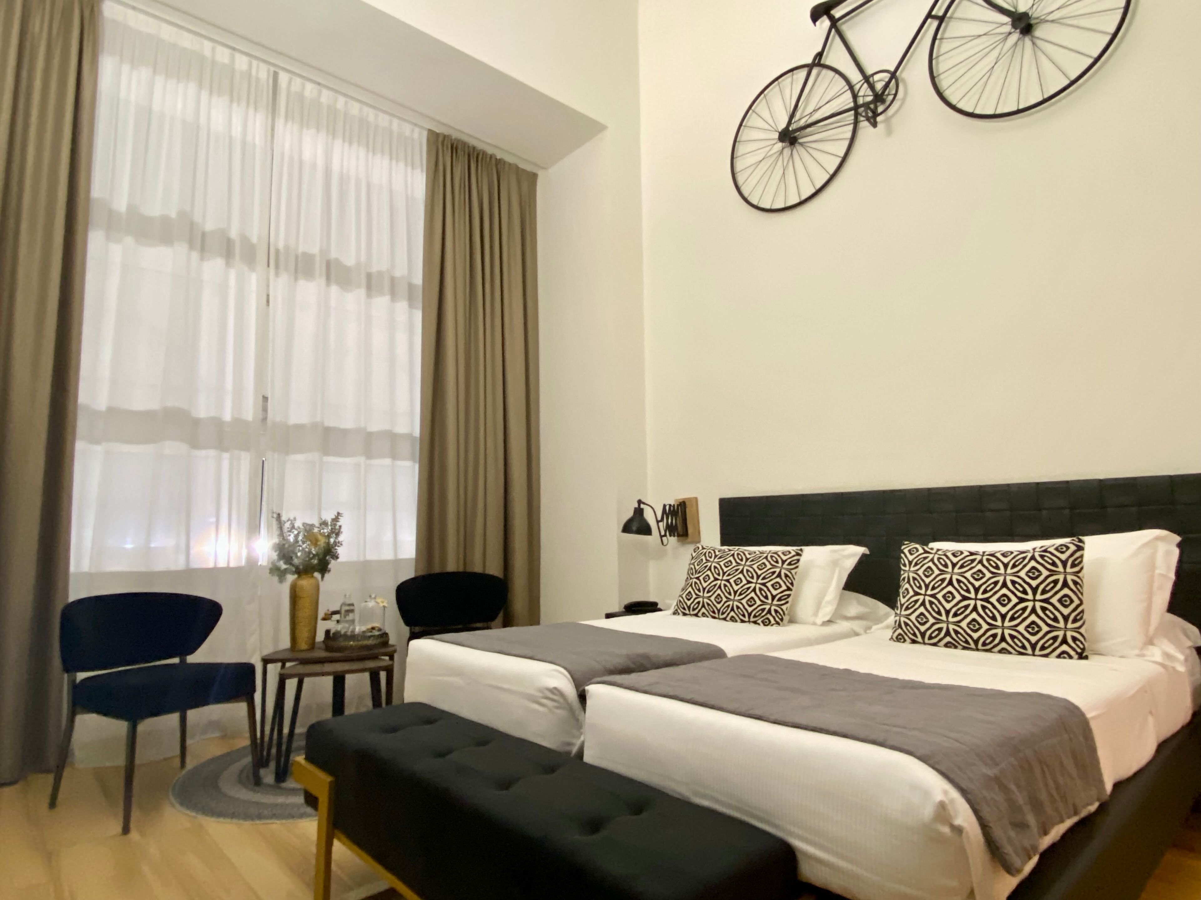 Ett hotellrum med en stor säng, två sängbord, en stol, ett litet bord med en vas och en cykel som hänger på väggen.