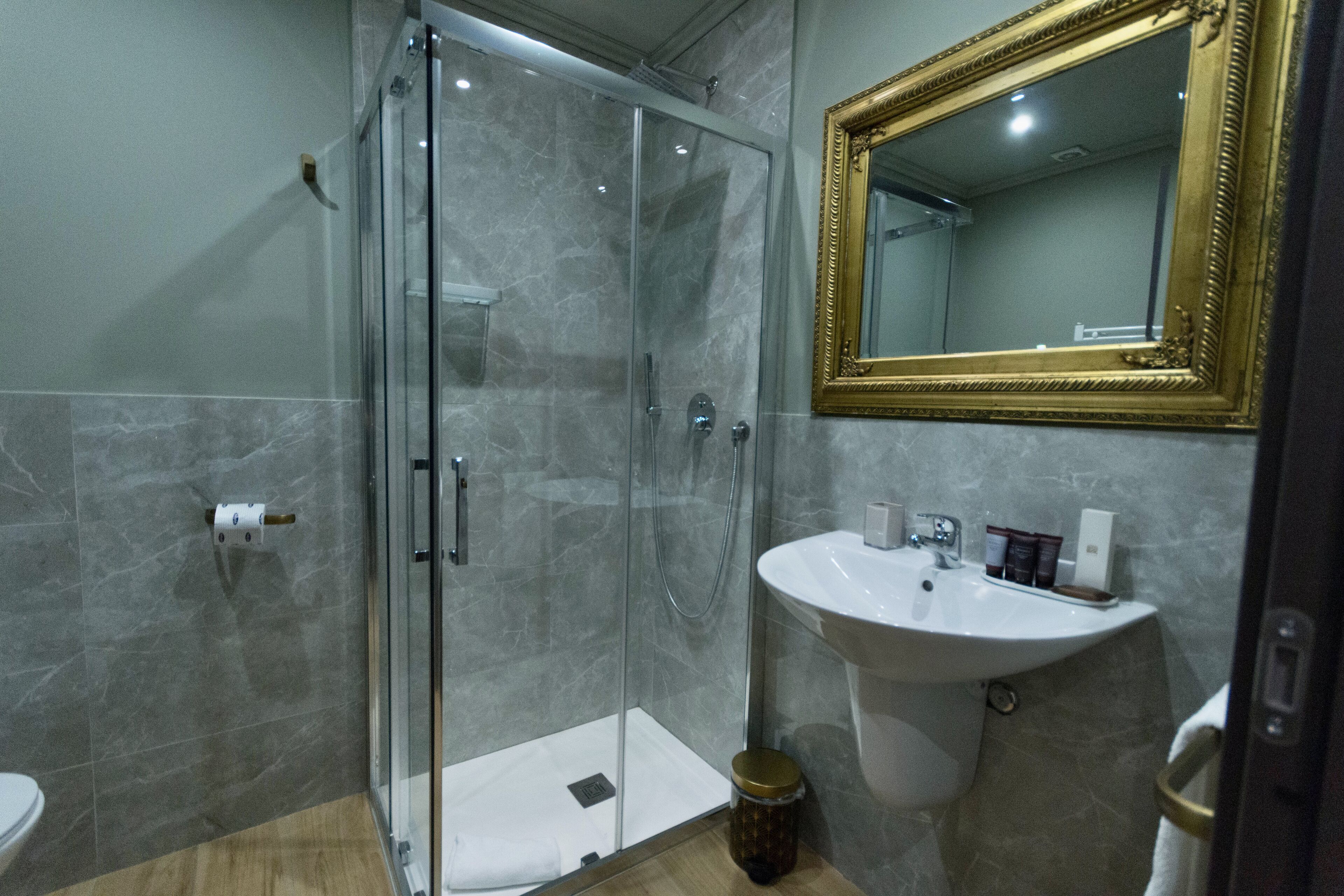 A bathroom with a glass shower enclosure, a white sink, and a mirror with a gold frame.