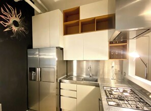 A modern kitchen with stainless steel appliances, including a refrigerator, stove, and sink. There are wooden shelves above the sink and a decorative sun on the wall. - INTOMILAN  Galleria Duomo - Boutique & Design Aparthotel (Milan)