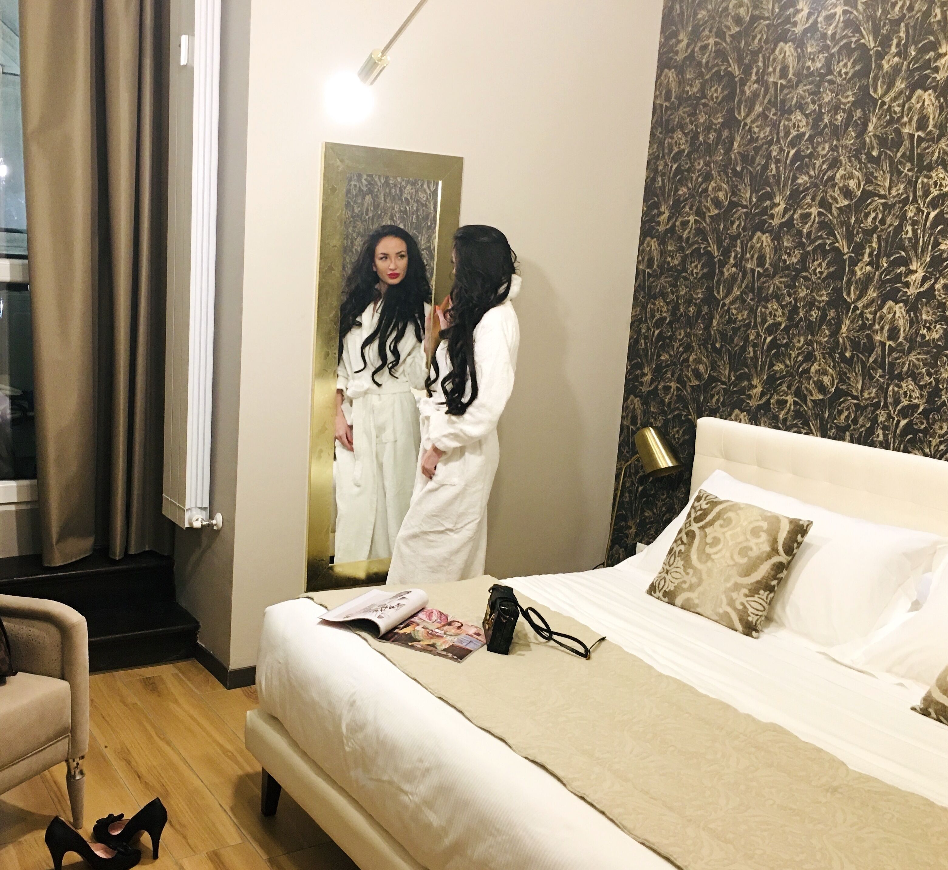 A hotel room with a bed, a mirror, a window with curtains, a lamp, and a magazine on the bed.