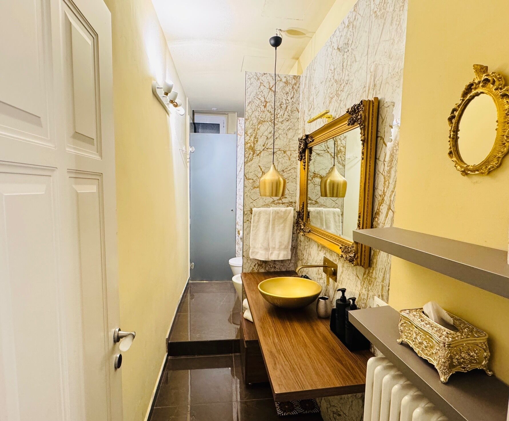 A narrow bathroom with a marble wall, a wooden vanity, a large yellow sink, and a gold-framed mirror.