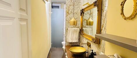 A narrow bathroom with a marble wall, a wooden vanity, a large yellow sink, and a gold-framed mirror.
