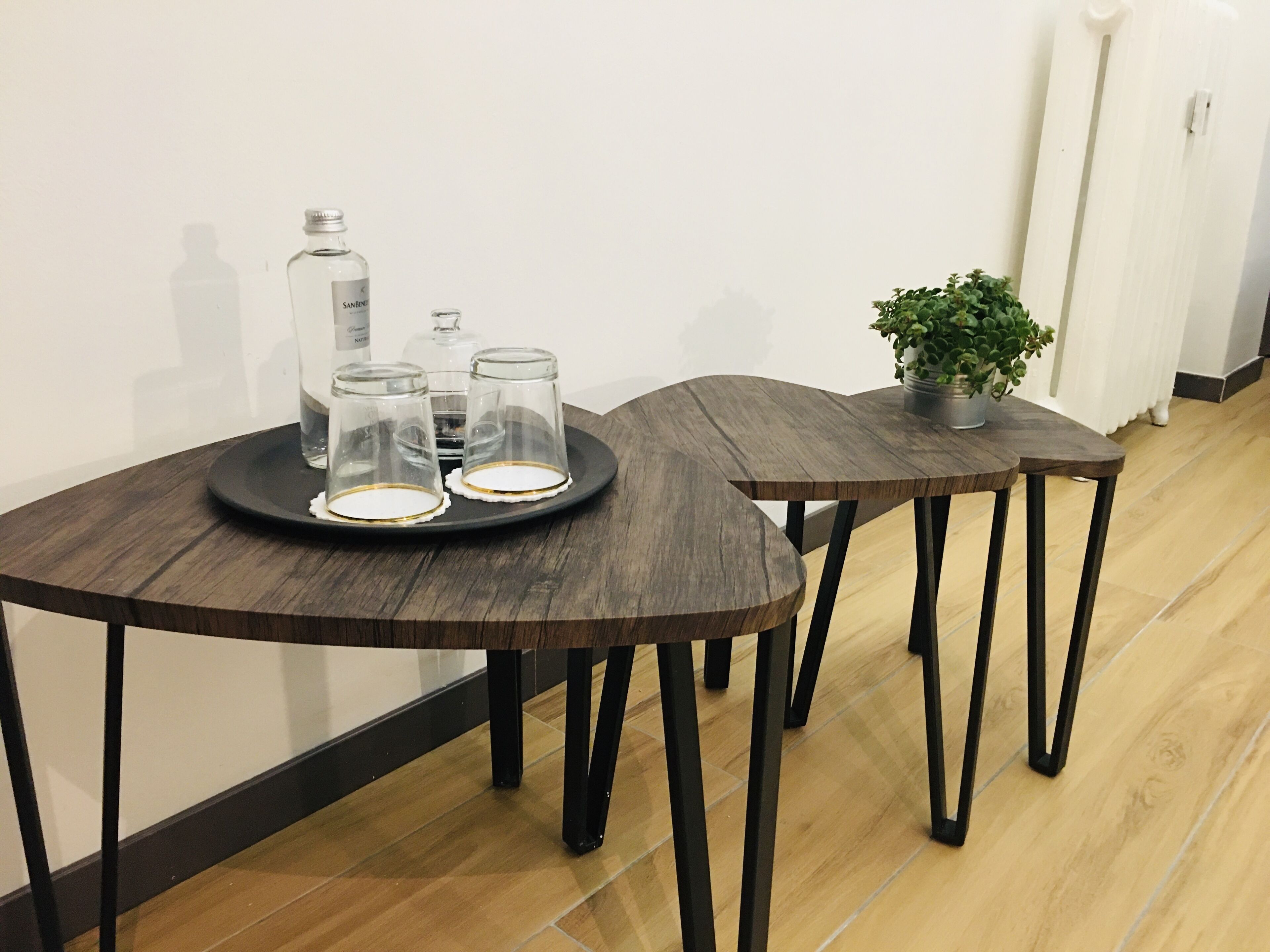 A room with two wooden tables, one with a tray holding three glass containers and a bottle, and another with a potted plant.