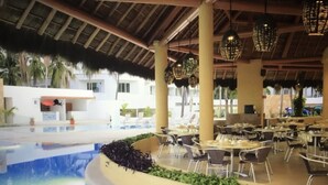 An indoor pool area with a thatched roof, surrounded by dining tables and chairs, and a pool with a water feature. - Ocean view 1King Bedroom/2BA, kitchen, private balcony, sofa bed, beachfront (Puerto Vallarta)