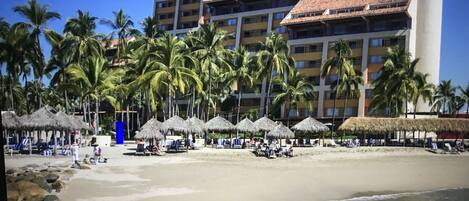 A beachfront hotel with multiple balconies, palm trees, and thatched umbrellas.
