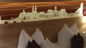 A hotel room with a bed, pillows, and a wall mural of a cityscape. - Hubertushöhe (Regensburg)