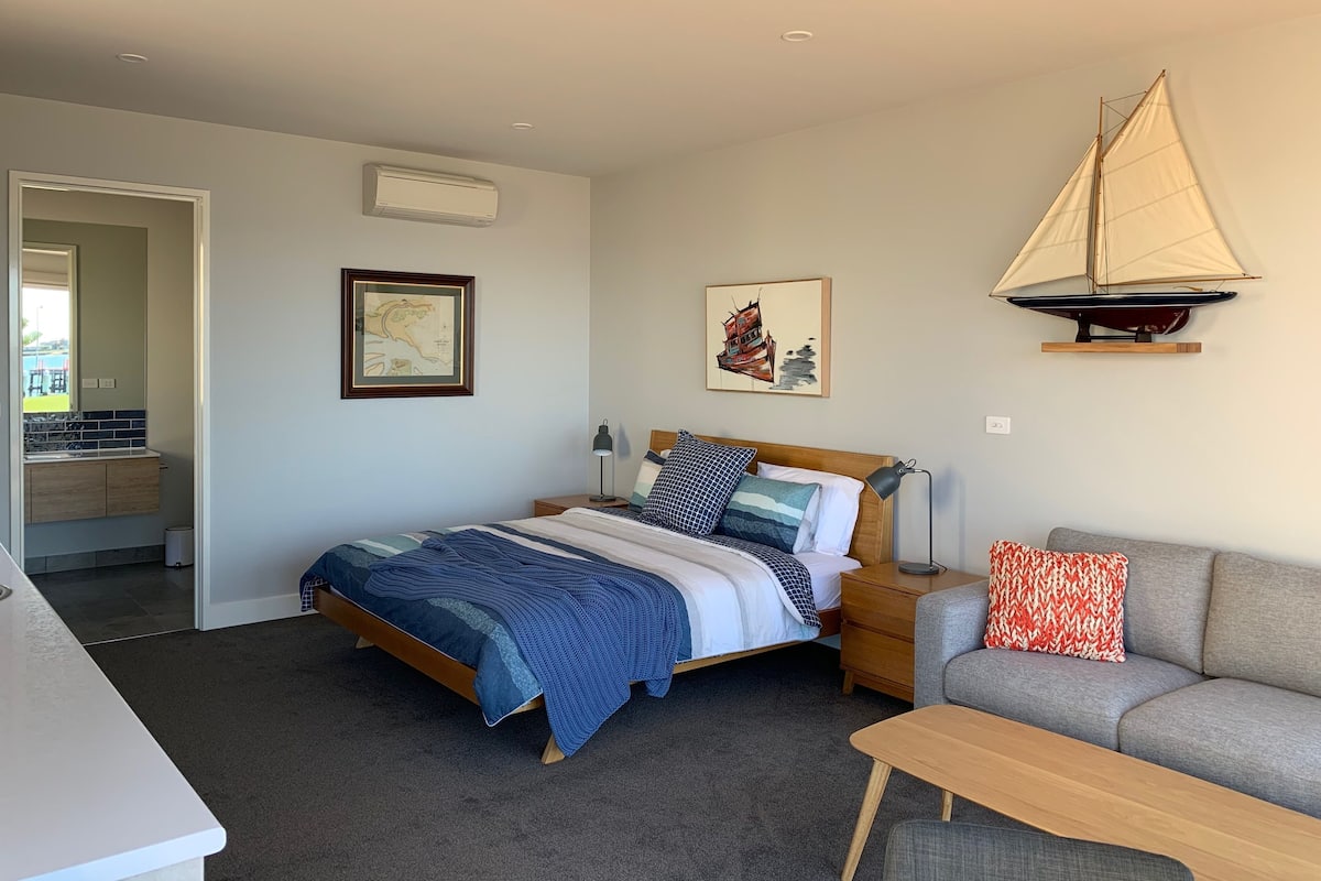 A bedroom with a bed, a nightstand, a desk, a chair, a sofa, and a sailboat decoration on the wall.