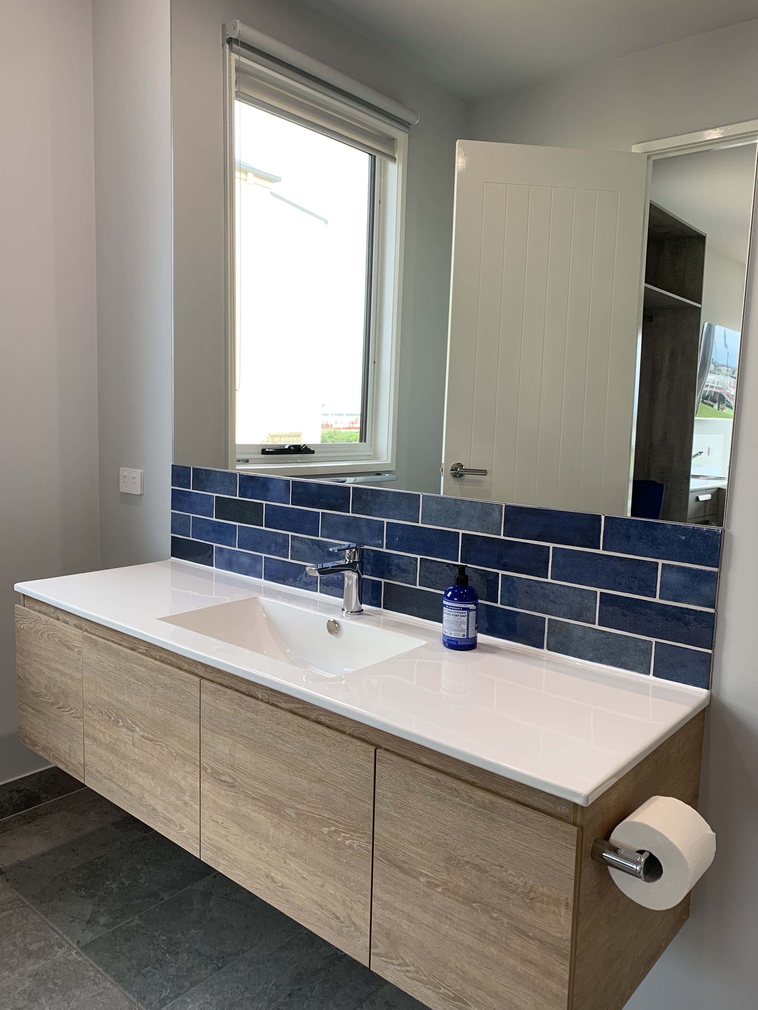 Une salle de bain avec un double lavabo, des meubles en bois, un pare-feu en carreaux bleus, une fenêtre et une porte.