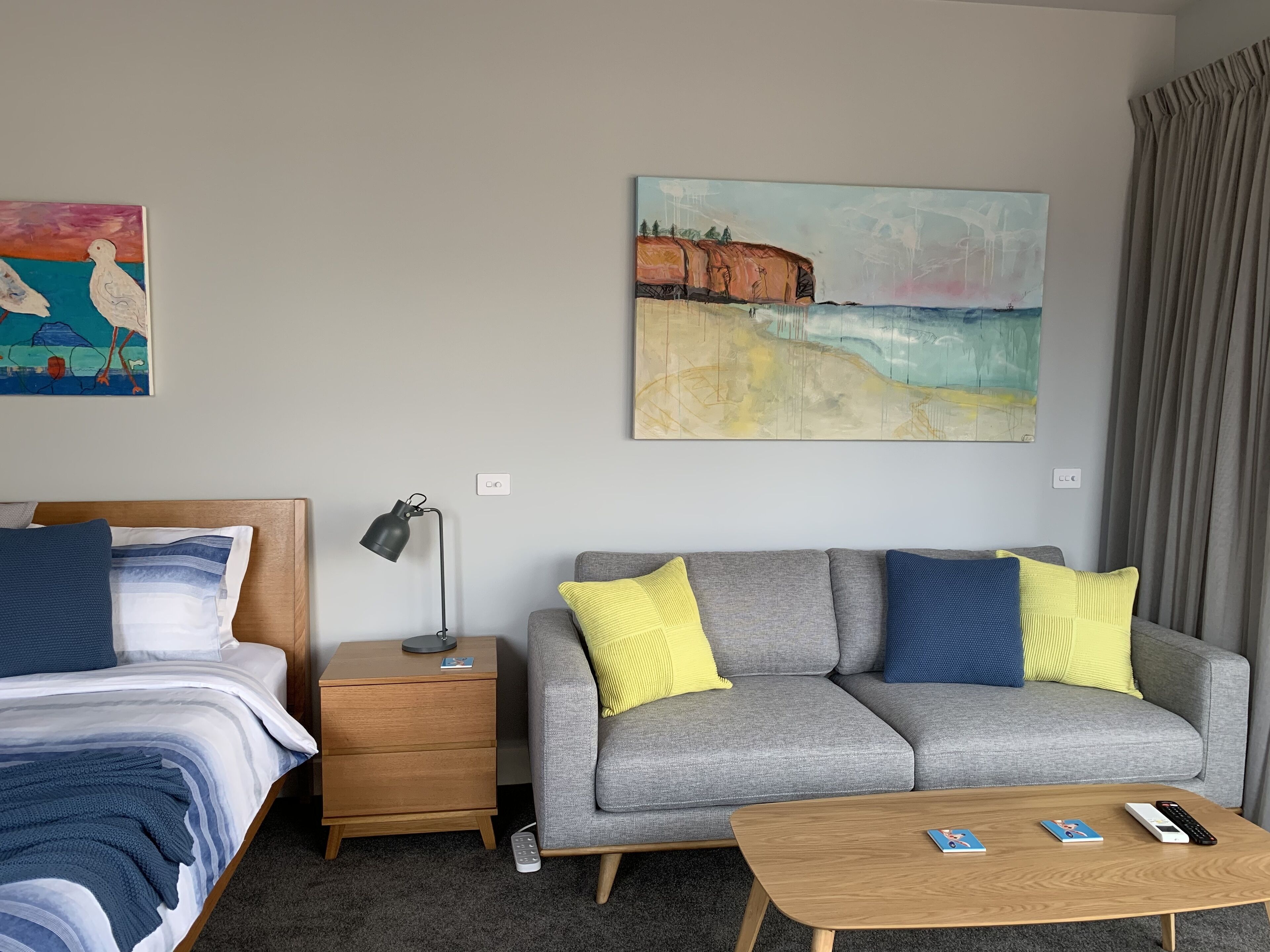 Ein Hotelzimmer mit einem Bett, einem grauen Sofa mit gelben und blauen Kissen, einem kleinen Nachttisch mit Lampe, einem Couchtisch mit Büchern und Fernbedienung sowie zwei Bildern an den Wänden.