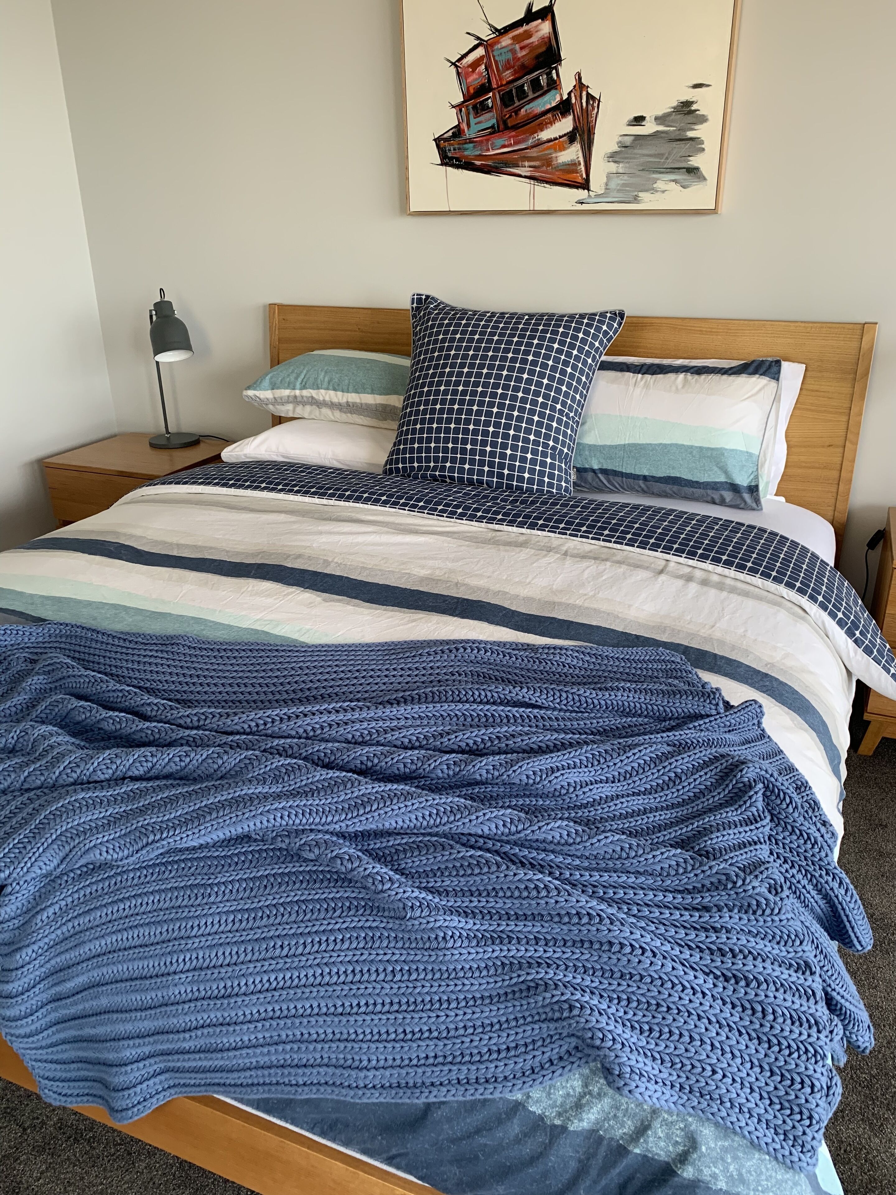 A neatly made bed with a blue knitted blanket, a patterned pillow, and a headboard with a painting above it.