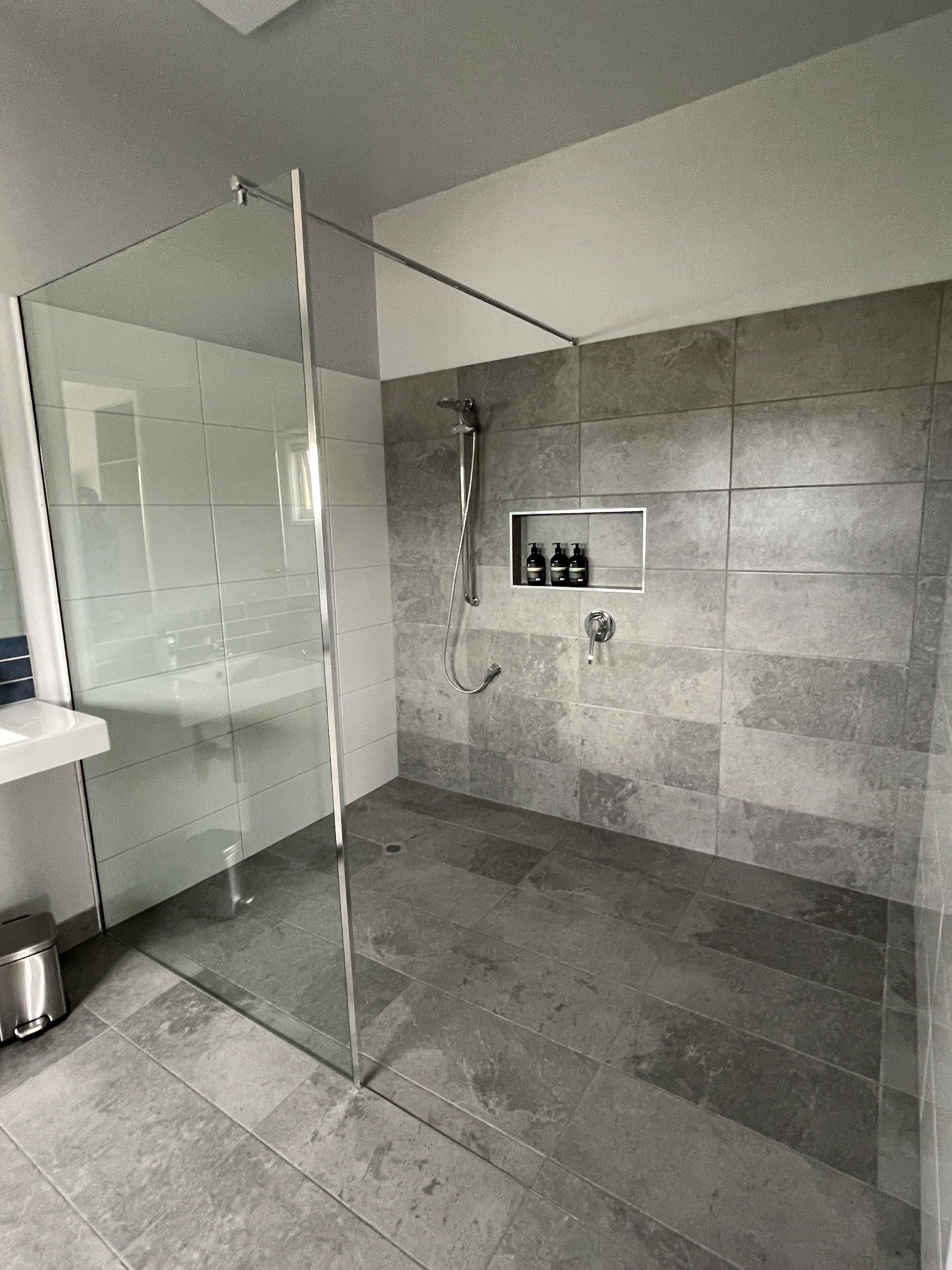 A modern bathroom with a glass shower enclosure, a built-in shelf with toiletries, and grey tiled walls and floor.