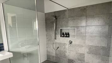 A modern bathroom with a glass shower enclosure, a built-in shelf with toiletries, and grey tiled walls and floor.