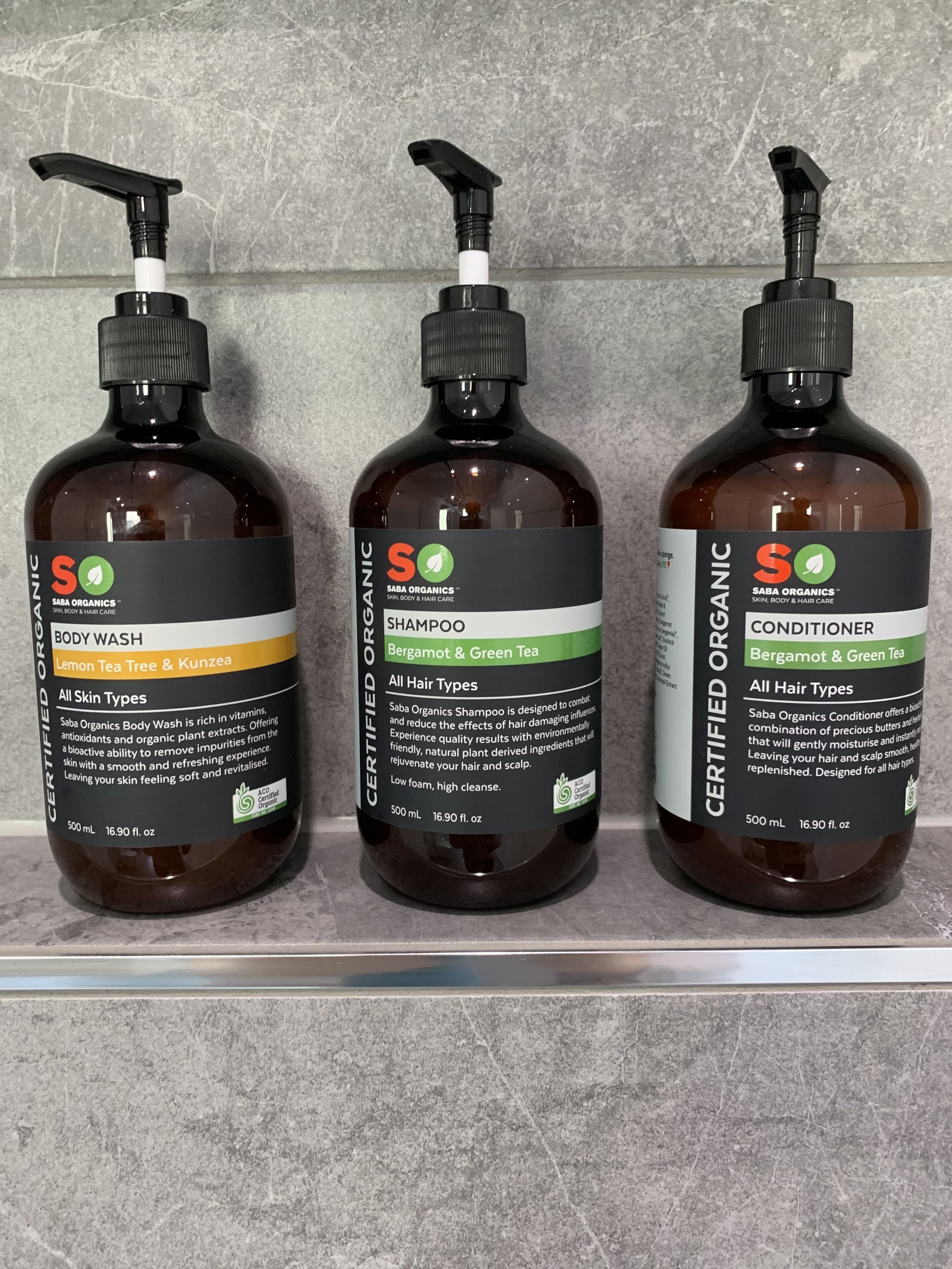 Three bottles of Saba Organics body wash, shampoo, and conditioner on a shelf.