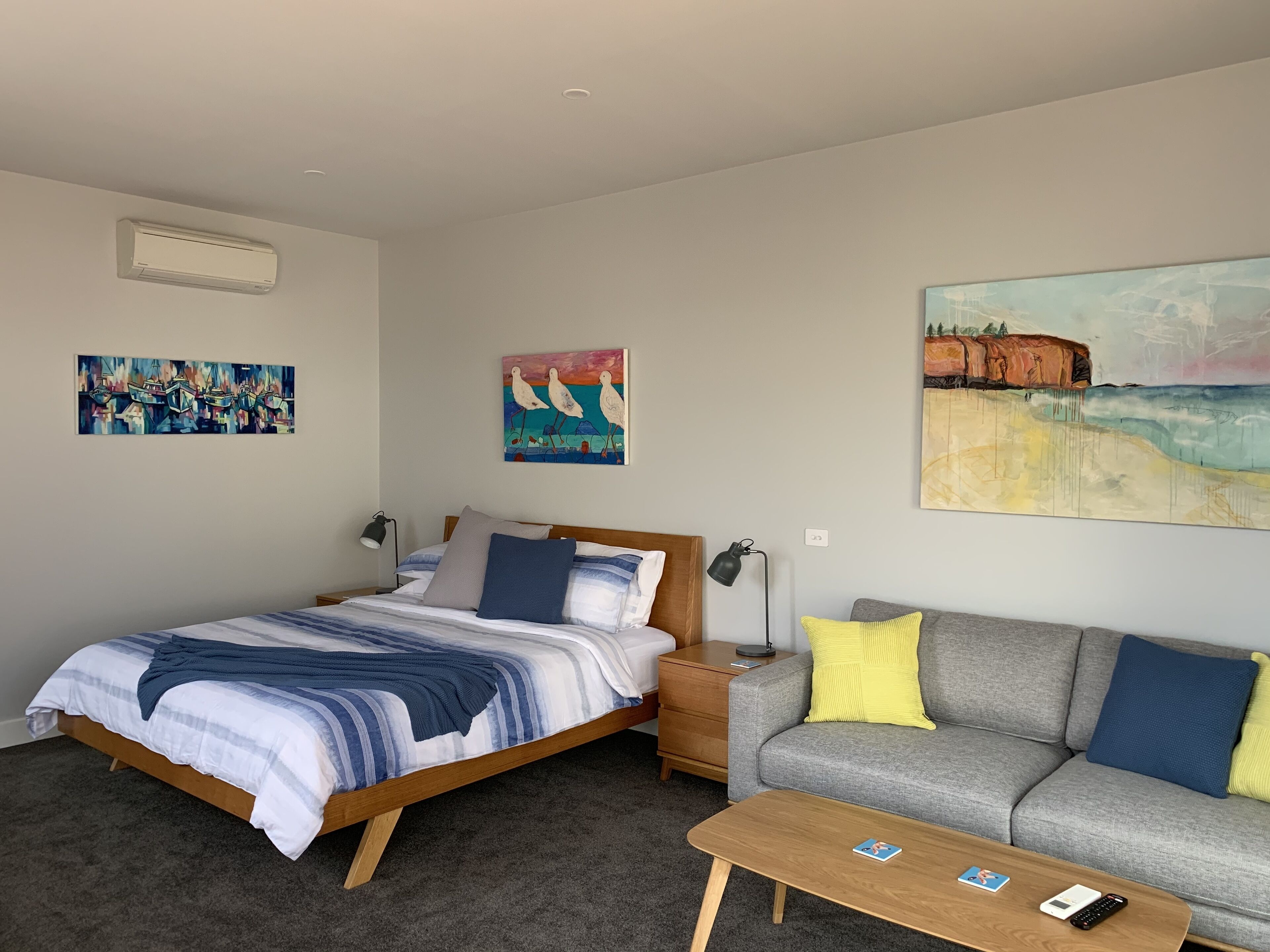 Une chambre d’hôtel moderne avec un lit, un canapé gris, une table basse en bois et des œuvres d’art accrochées aux murs.