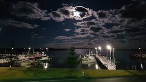 Una marina de noche con barcos amarrados, un muelle que se adentra en el agua y un puente iluminado.