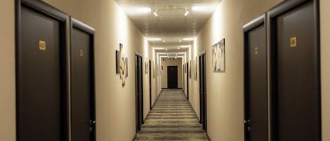 A hotel hallway with doors on both sides, a patterned carpet, and framed pictures on the walls.