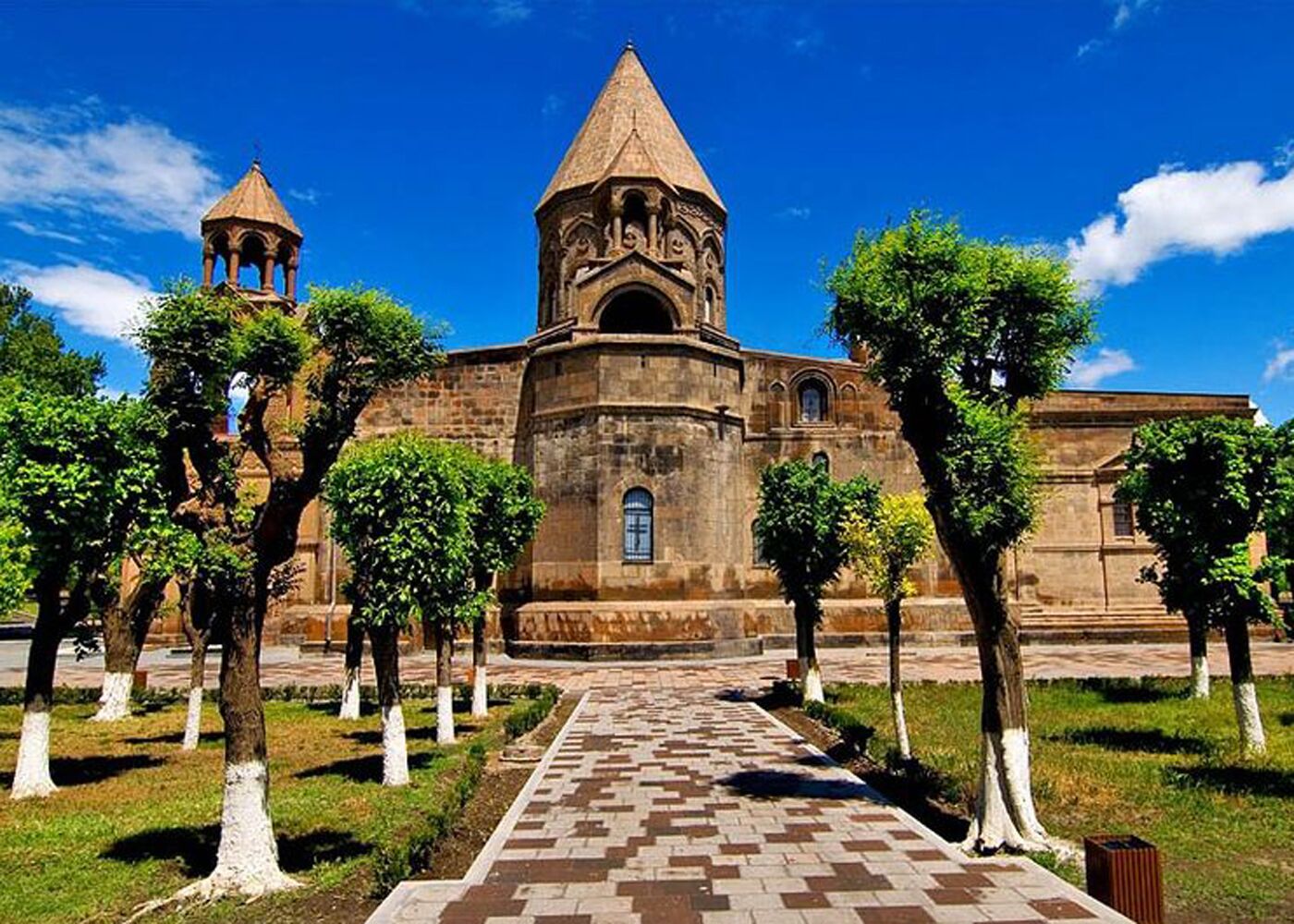 Uma igreja histórica com cúpula central e um campanário, rodeada por um jardim simétrico com árvores podadas e um caminho pavimentado.