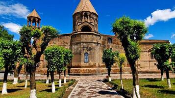 Uma igreja histórica com cúpula central e um campanário, rodeada por um jardim simétrico com árvores podadas e um caminho pavimentado.