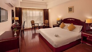 A hotel room with a large bed, two bedside tables with lamps, a desk, a chair, a small table, and a ceiling fan. - Mayur aaditya resort (Dharwad)