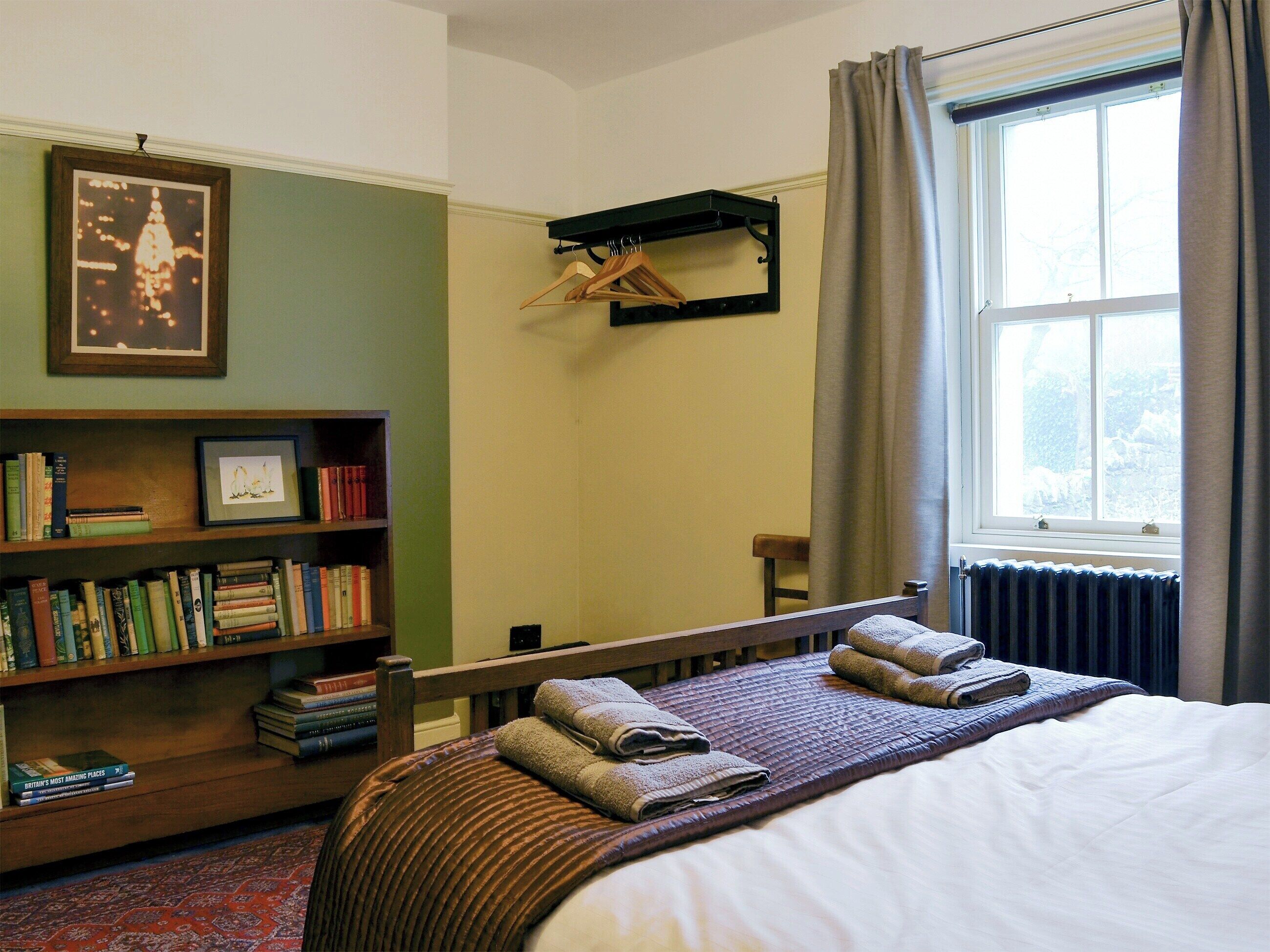 Une chambre à coucher bien rangée, avec un grand lit, une bibliothèque remplie de livres, un tableau encadré au mur et une fenêtre avec des rideaux.