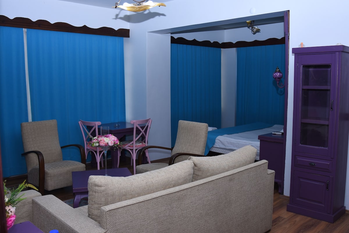 Ein Zimmer mit einem Bett, zwei Stühlen, einem kleinen Tisch mit einer Vase und einem violetten Schrank.