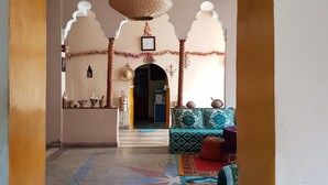 A room with a large wooden door, a hanging lamp, a sofa with patterned cushions, and a red rug. - Gîte Kasbah La palmeraie (Skoura)