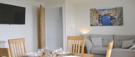 A dining area with a wooden table set for four, a grey sofa, and a painting of a coastal landscape.