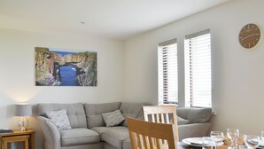Uma sala de estar moderna com um sofá cinzento, mesa de jantar de madeira para quatro pessoas e um quadro com paisagem costeira na parede.