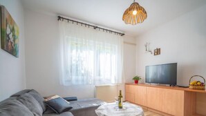 Un salon moderne avec un canapé gris, une petite table ronde sur laquelle se trouve une bouteille, un téléviseur posé sur un meuble en bois et une fenêtre ornée de rideaux légers.