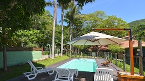 Una terraza de madera con piscina, sillas de descanso blancas y un amplio paraguas.