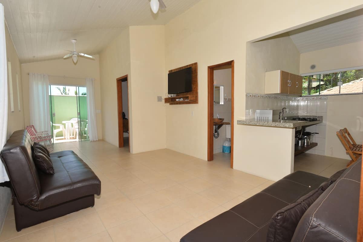 A spacious living area with a brown leather sofa, a television mounted on the wall, and a kitchen area with a countertop and sink.