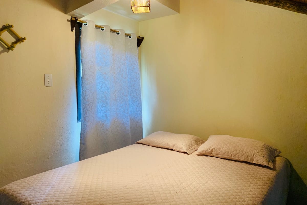 A single bed with a quilted mattress and two pillows in a room with plain walls and a curtain.