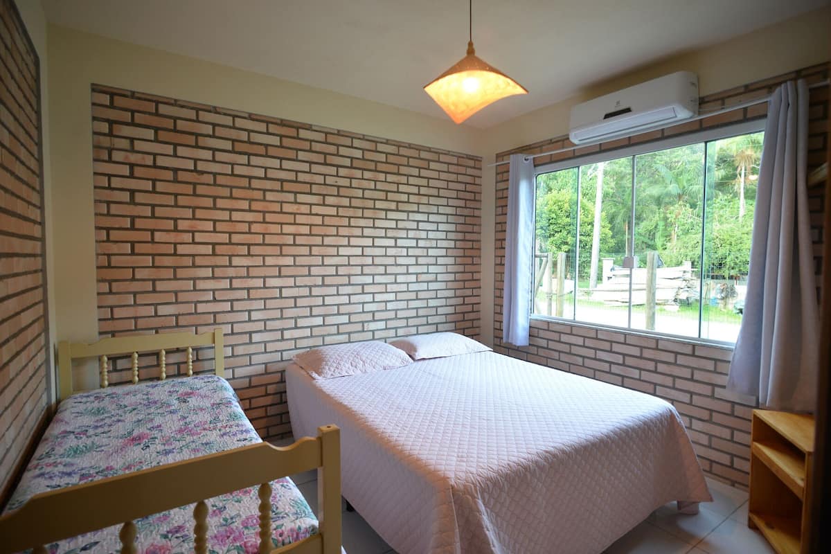 A room with a brick wall, a bed with a quilted cover, a wooden chair, a window with curtains, and an air conditioning unit.