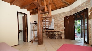Una habitación con escalera de madera, pared de ladrillo, zona de comedor con sillas y cama con colcha rosa.