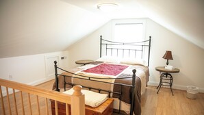 A bedroom with a sloped ceiling, a bed with a metal frame, a wooden chest of drawers, a small round table with a lamp, and a waste basket.