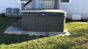 A hot tub in front of a house with a wooden deck and two chairs.