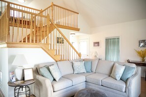 Un salon avec un escalier en bois, un canapé d’angle gris et une petite table avec une lampe.