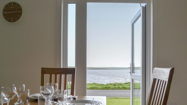 Una mesa de comedor dispuesta con platos, vasos y cubiertos, frente a una ventana que ofrece vistas a un cuerpo de agua y un paisaje lejano.