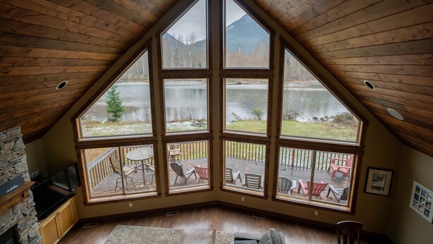 Ein Zimmer mit großem Fenster, das auf einen See und Berge blickt, mit Holzboden, Kamin aus Stein und einer Steinwand.