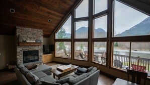 Ein geräumiges Wohnzimmer mit Kamin aus Stein, großen Fenstern mit Blick auf einen See und Berge sowie einer gemütlichen Sitzecke.