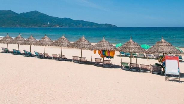 Ein Strand mit Liegestühlen, strohgedeckten Sonnenschirmen, einem Rettungsschwimmerstand und einem Berg im Hintergrund.