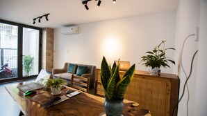 Ein modernes Wohnzimmer mit Couchtisch aus Holz, einem Laptop, einem Smartphone und einer Topfpflanze. Es gibt eine Couch mit Kissen und ein Fenster mit Blick ins Freie.