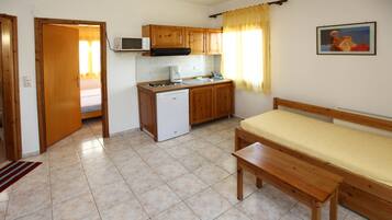 Cocina compacta con armarios de madera, microondas, lavamanos y un refrigerador pequeño. Hay un banco de madera y una cama en la habitación contigua.