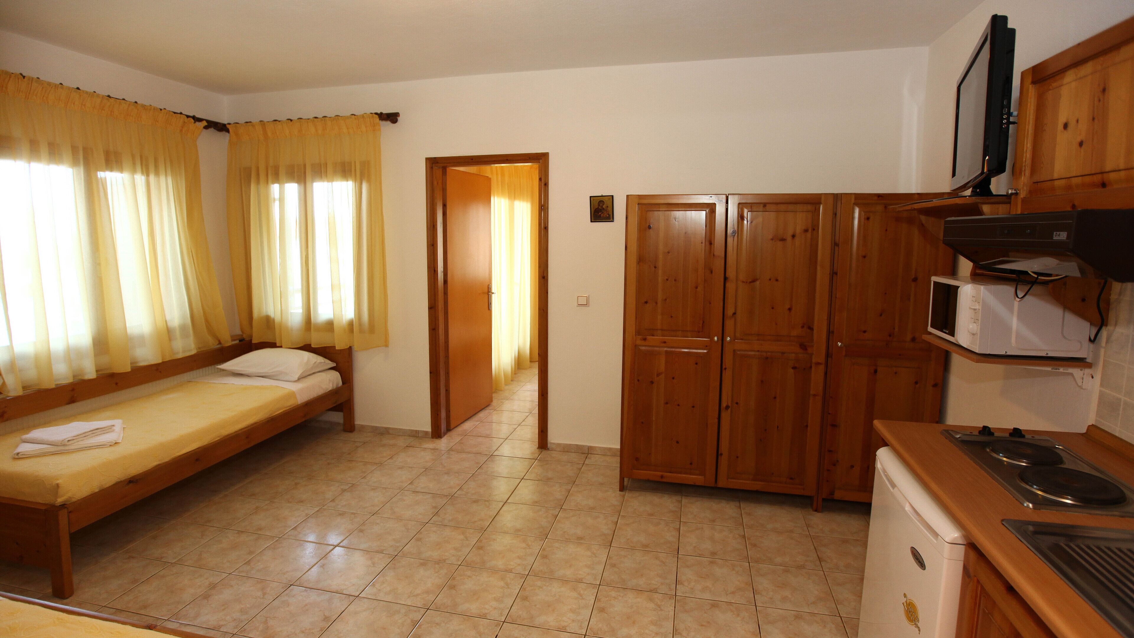 A small, compact room with a bed, kitchenette, and wooden wardrobe.