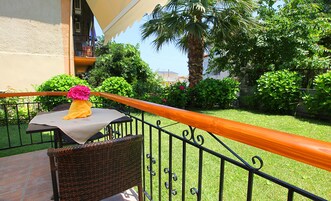 Un balcón con una silla de mimbre, una mesita con un mantel amarillo y un arreglo floral rosa, y vistas a un jardín cuidado con diversas plantas y una palmera.