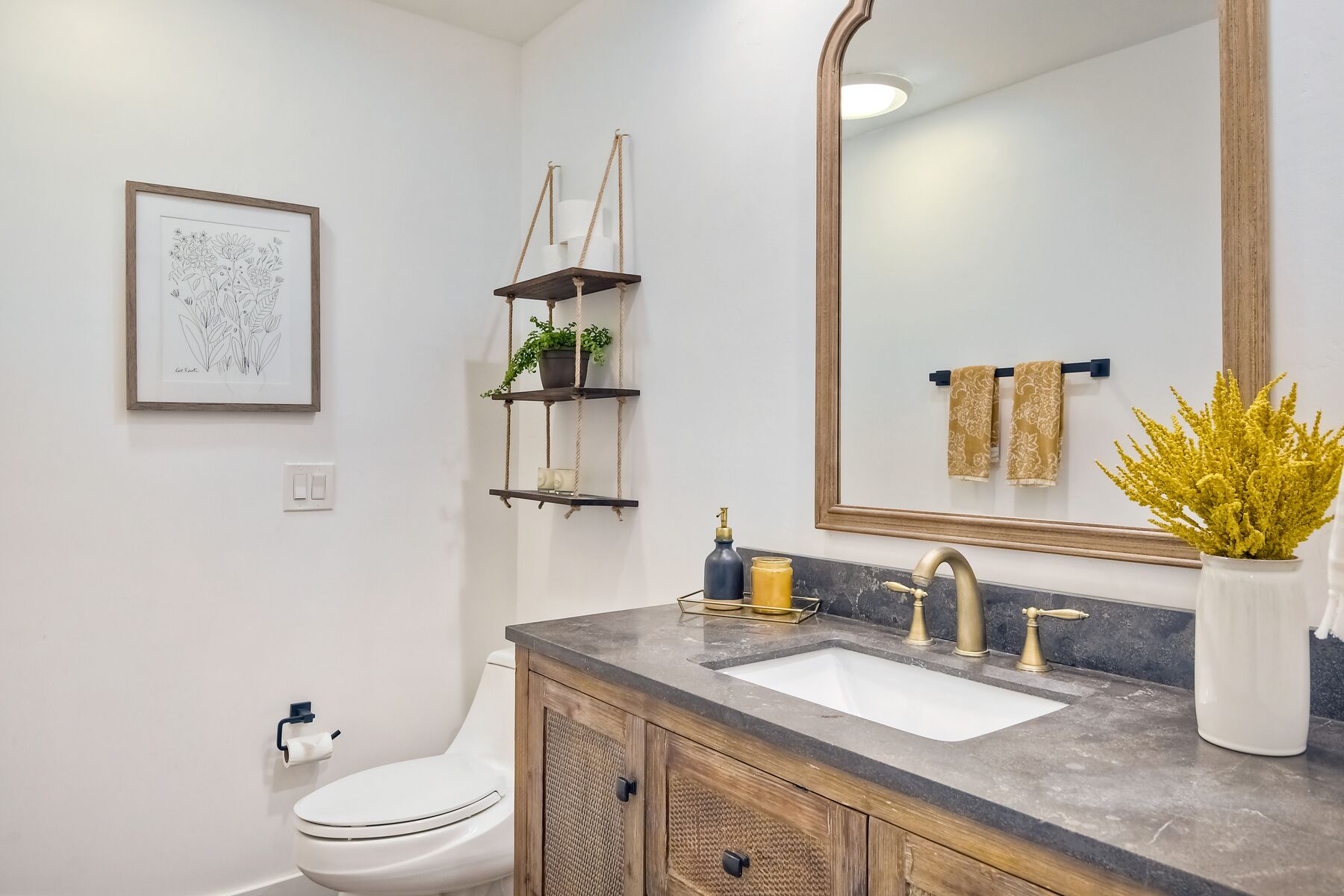 A bathroom with a white toilet, a sink with a marble countertop, a large mirror, a framed botanical illustration, and a plant on the counter.